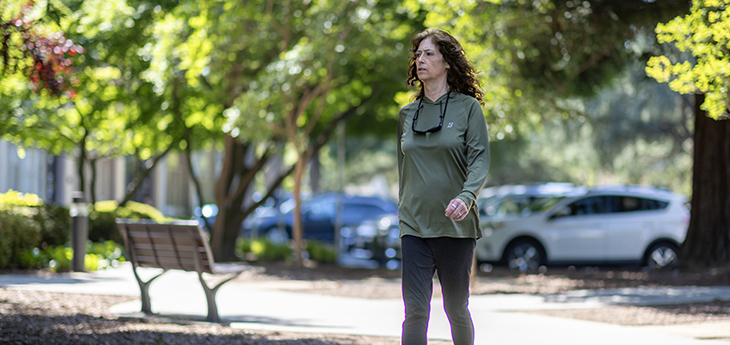 Woman with dark hair, wearing green sweatshirt and navy pants, walks down sidewalk with trees and park bench in background.